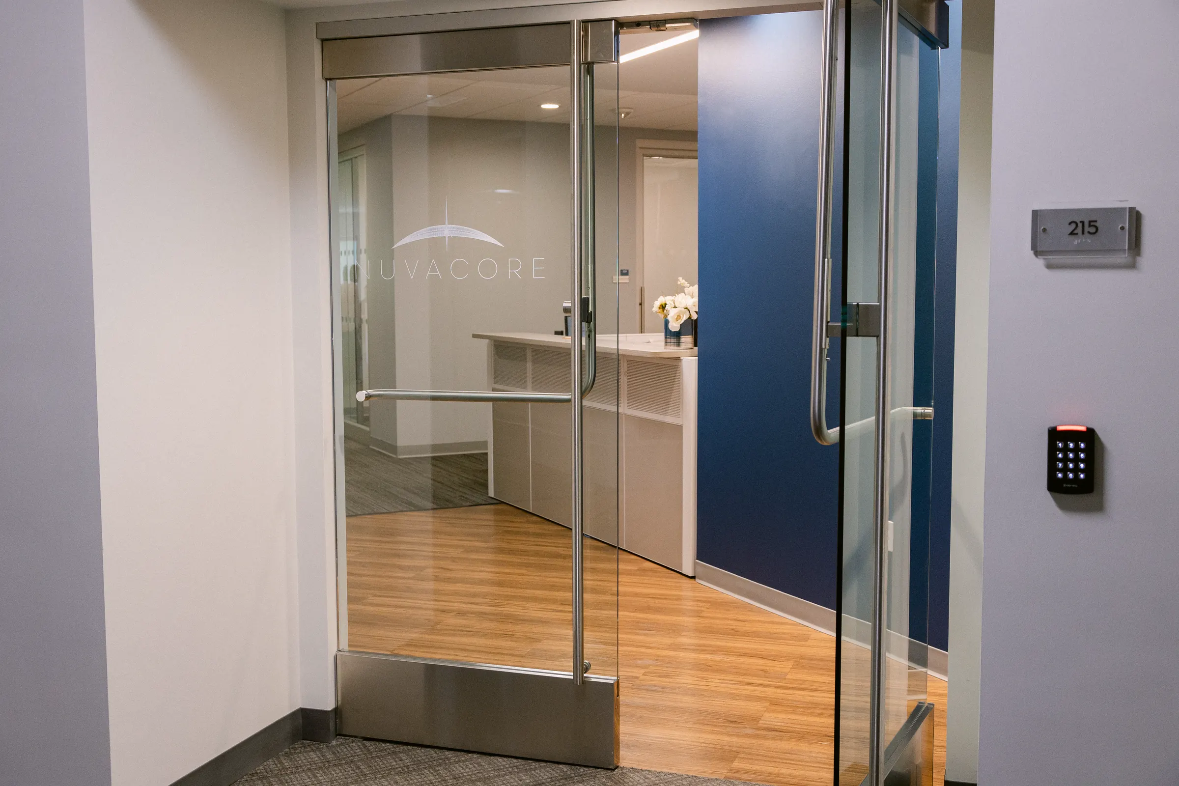 The glass door of NUVACORE\u2019s Santa Clara office, with the etched NUVACORE mark and reception behind it